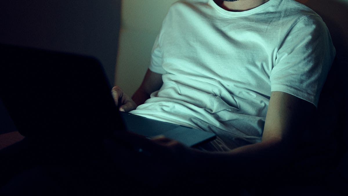 Man in a white t-shirt browsing alone in the dark, representing signs of porn addiction and isolation.