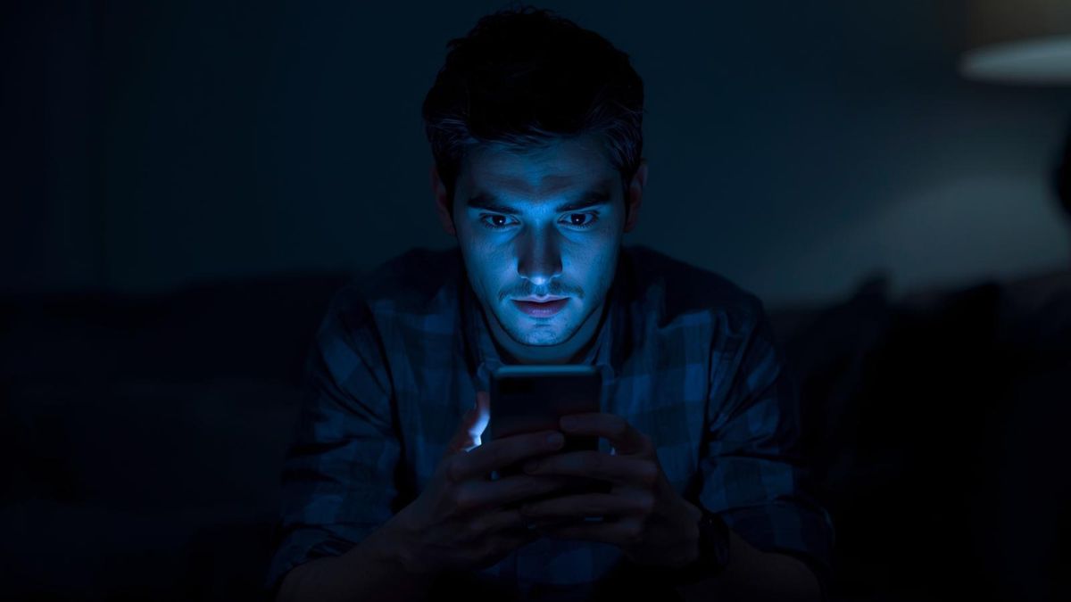 A man sitting alone in the dark illuminated by a smartphone screen, representing social media addiction.
