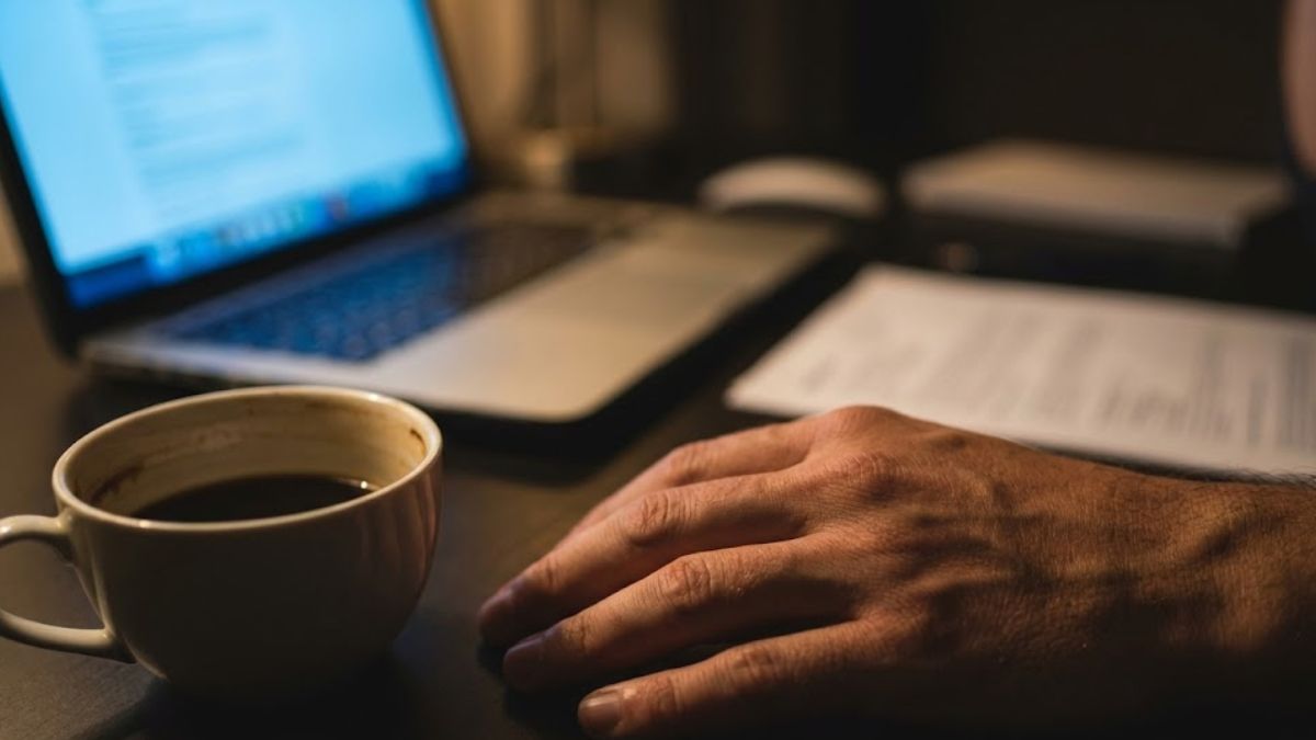 A hand resting near a coffee cup late at night with a laptop in the background, representing insomnia caused by stress.