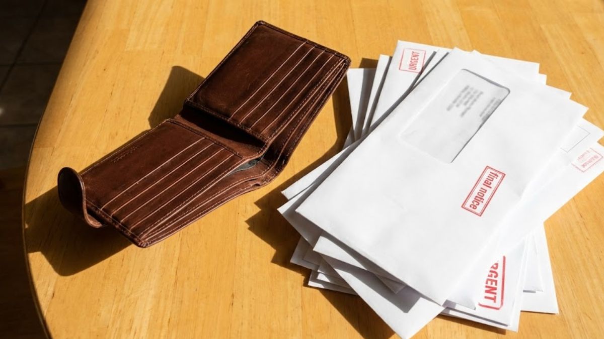 An empty wallet on a table next to a pile of unopened bill envelopes.