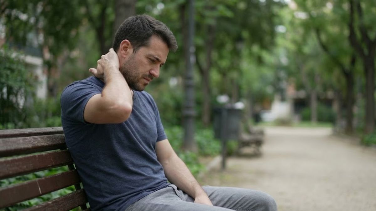 A man sitting on a park bench rubbing the back of his neck, showing physical signs of tension or stress.