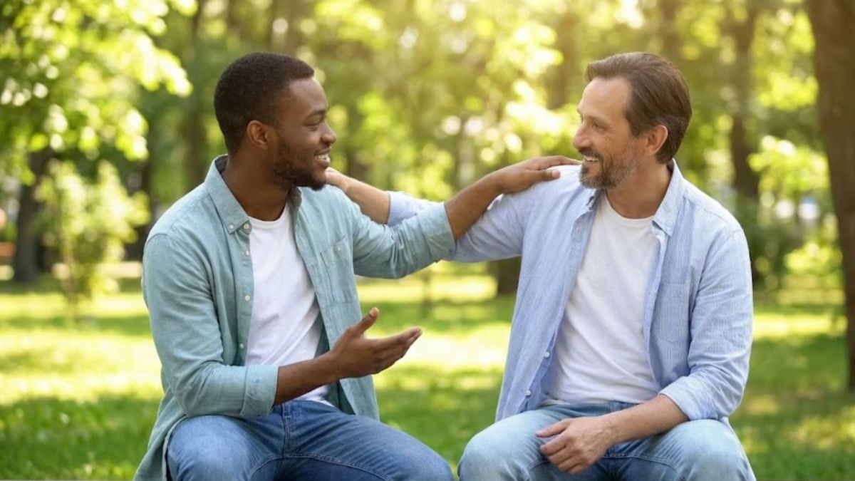 Two men sitting on a park bench, having a friendly and supportive conversation, with one man's hand on the other's shoulder.