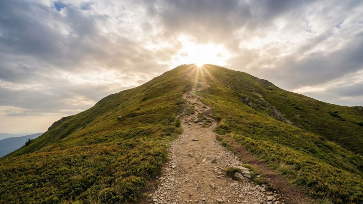 A view from a steep hiking trail, looking up towards a mountain summit where the sun is breaking through the clouds.