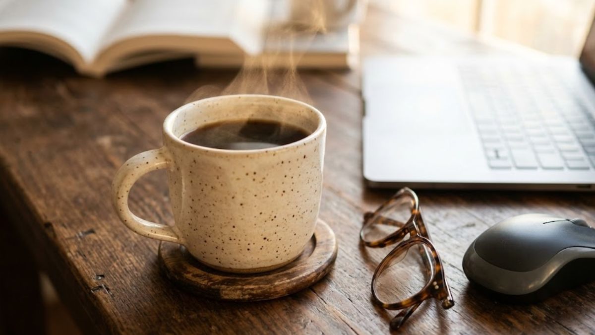 A warm, steaming cup of coffee next to a computer mouse on a wooden desk.